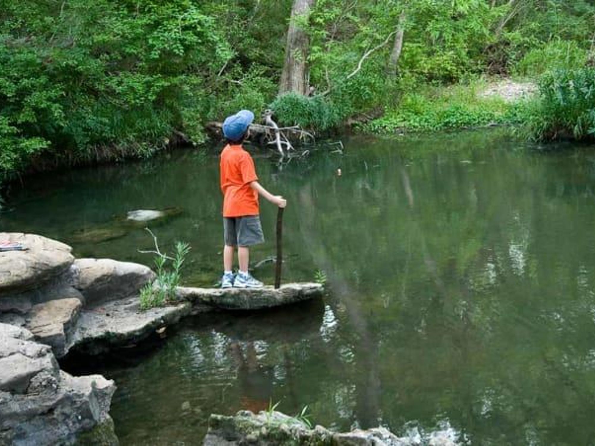 A child stands on a rock formation, holding a walking stick and looking over a river