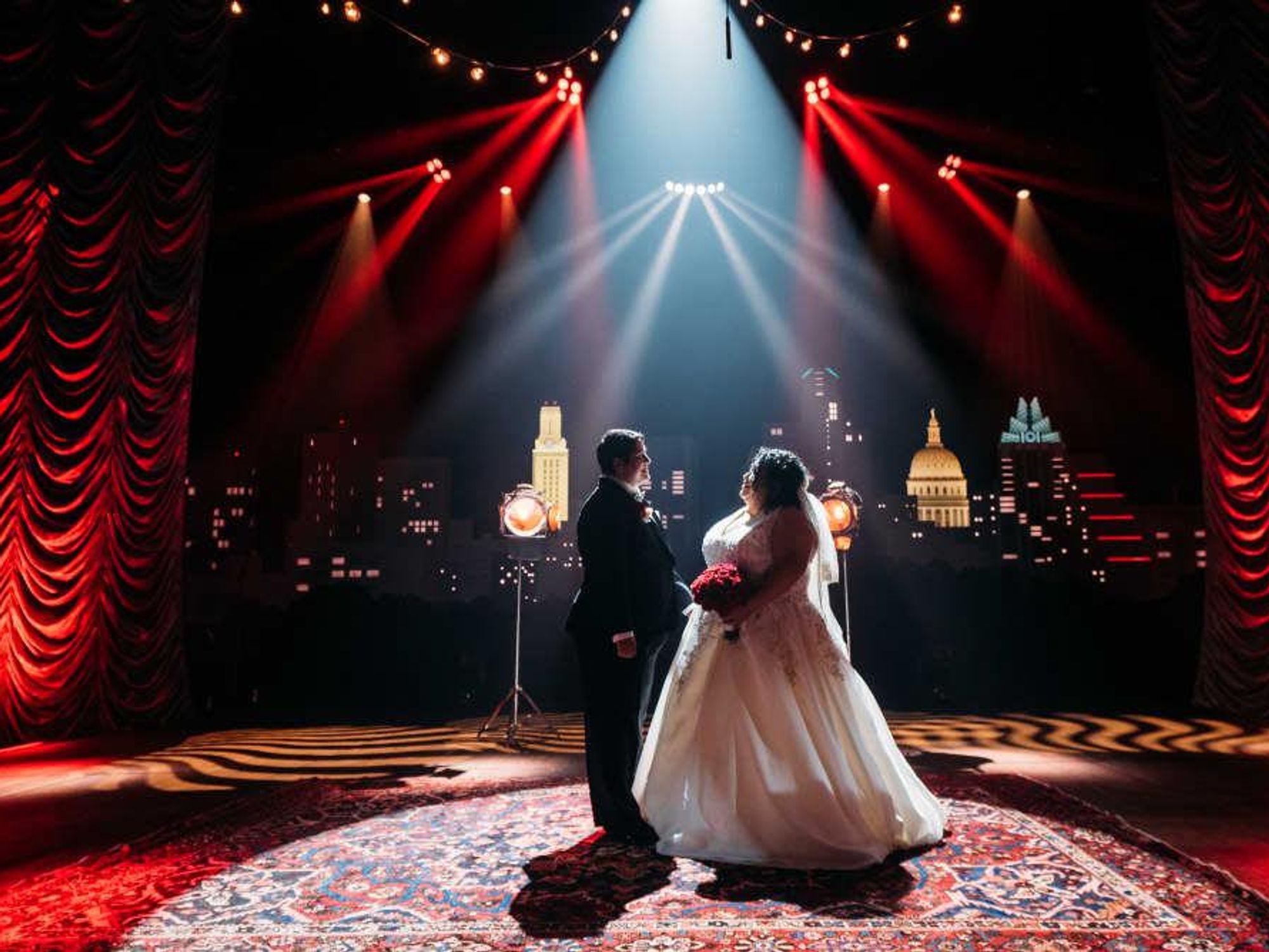 A couple stands smiling at each other in a wedding dress and tuxedo, in front of red curtains and the recognizable skyline backdrop at Austin City Limits Live.