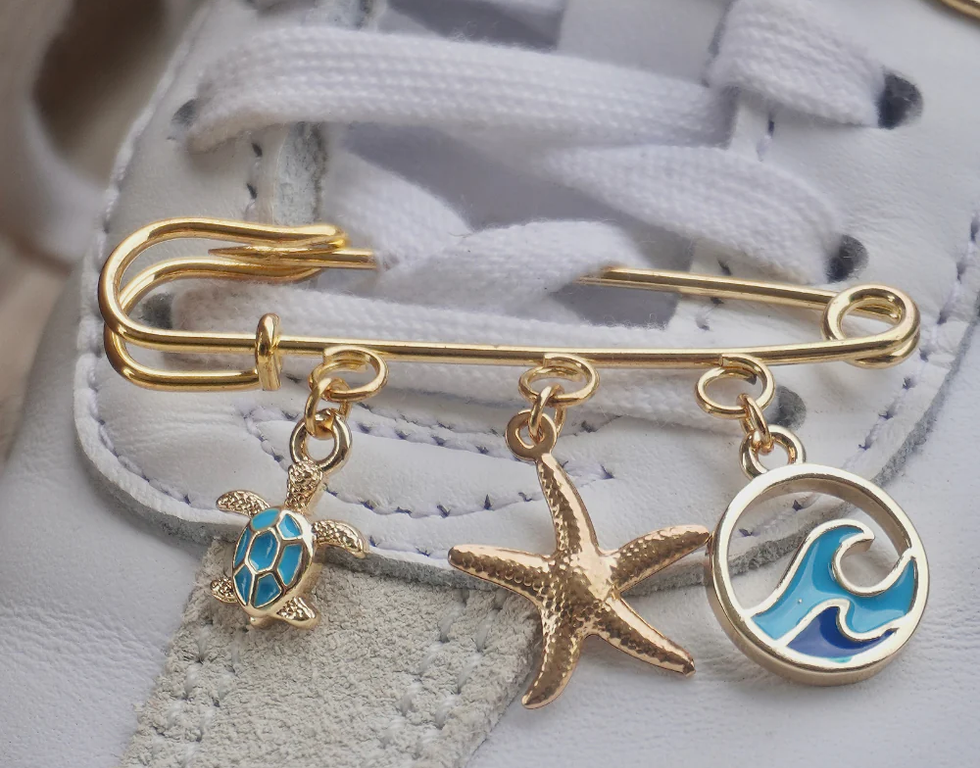 A gold safety pin with ocean-inspired charms, including a turtle, a starfish, and a blue wave, pinned to a shoelace on top of a shoe.