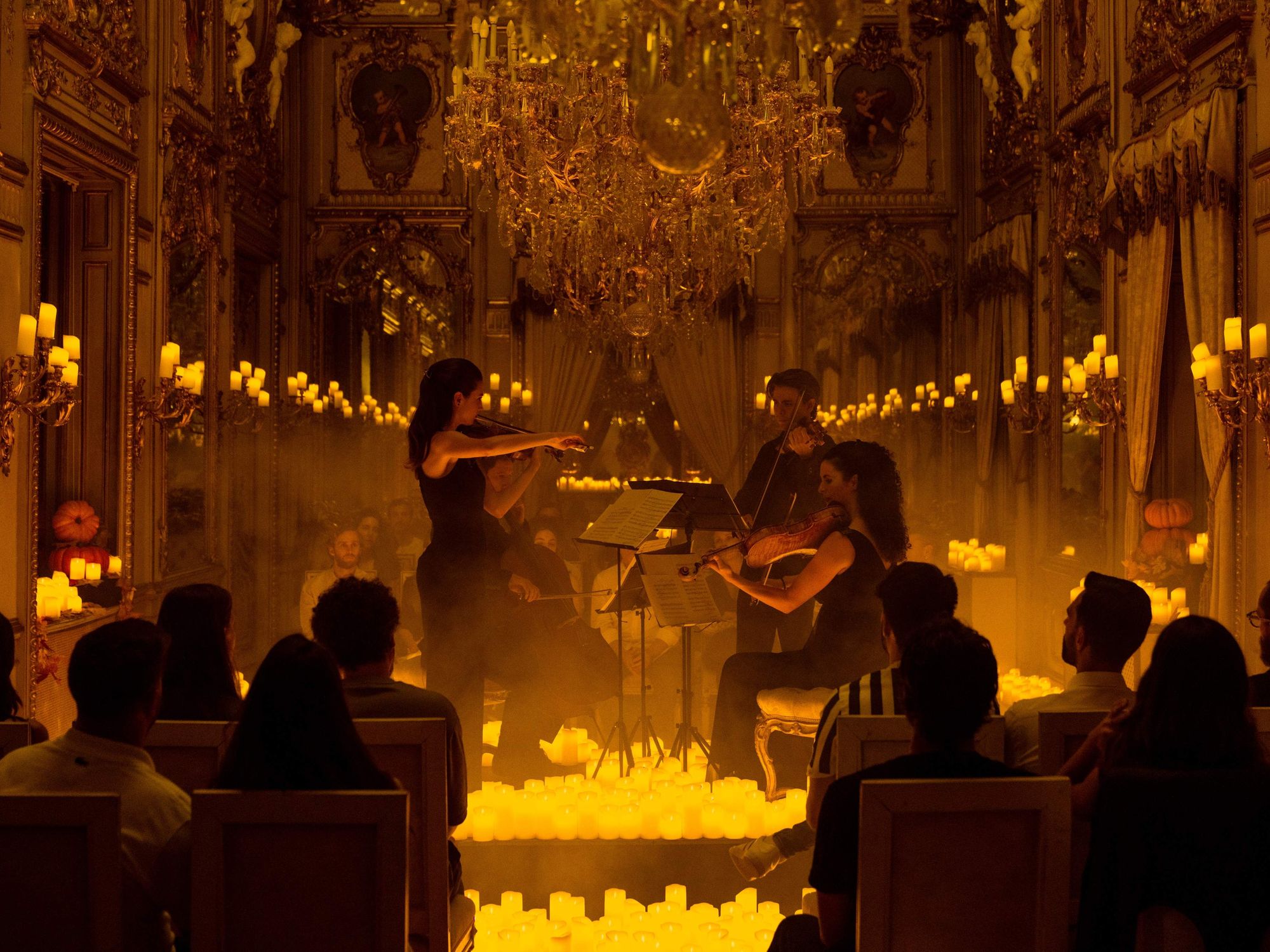 A group of violinists play for an audience, surrounded by candles.