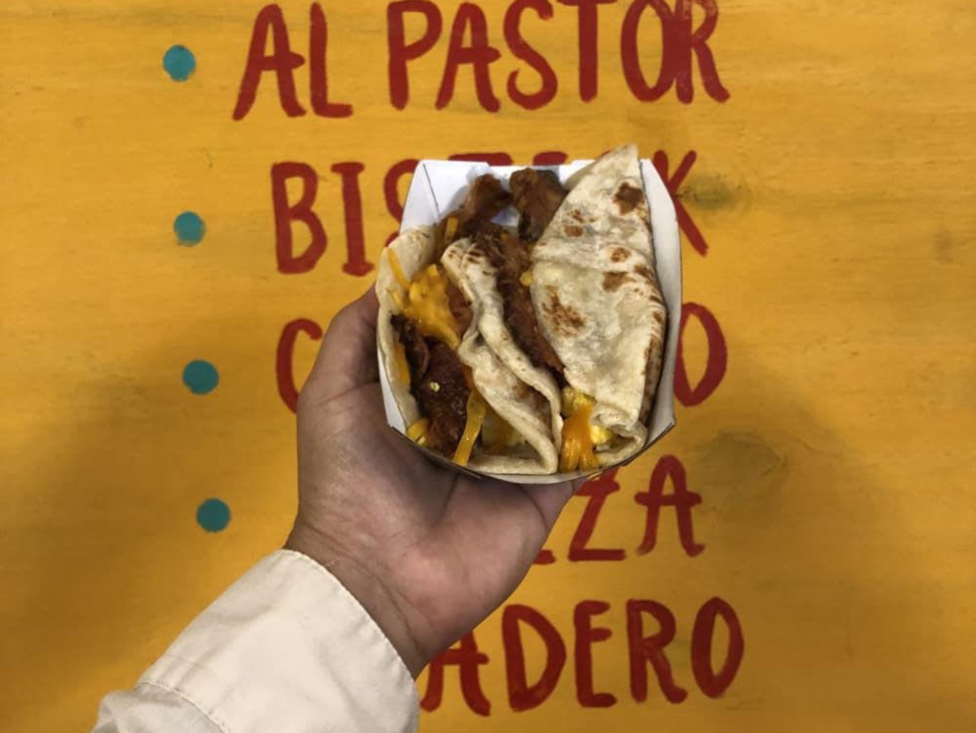 A hand displays a taco in front of a handwritten menu.