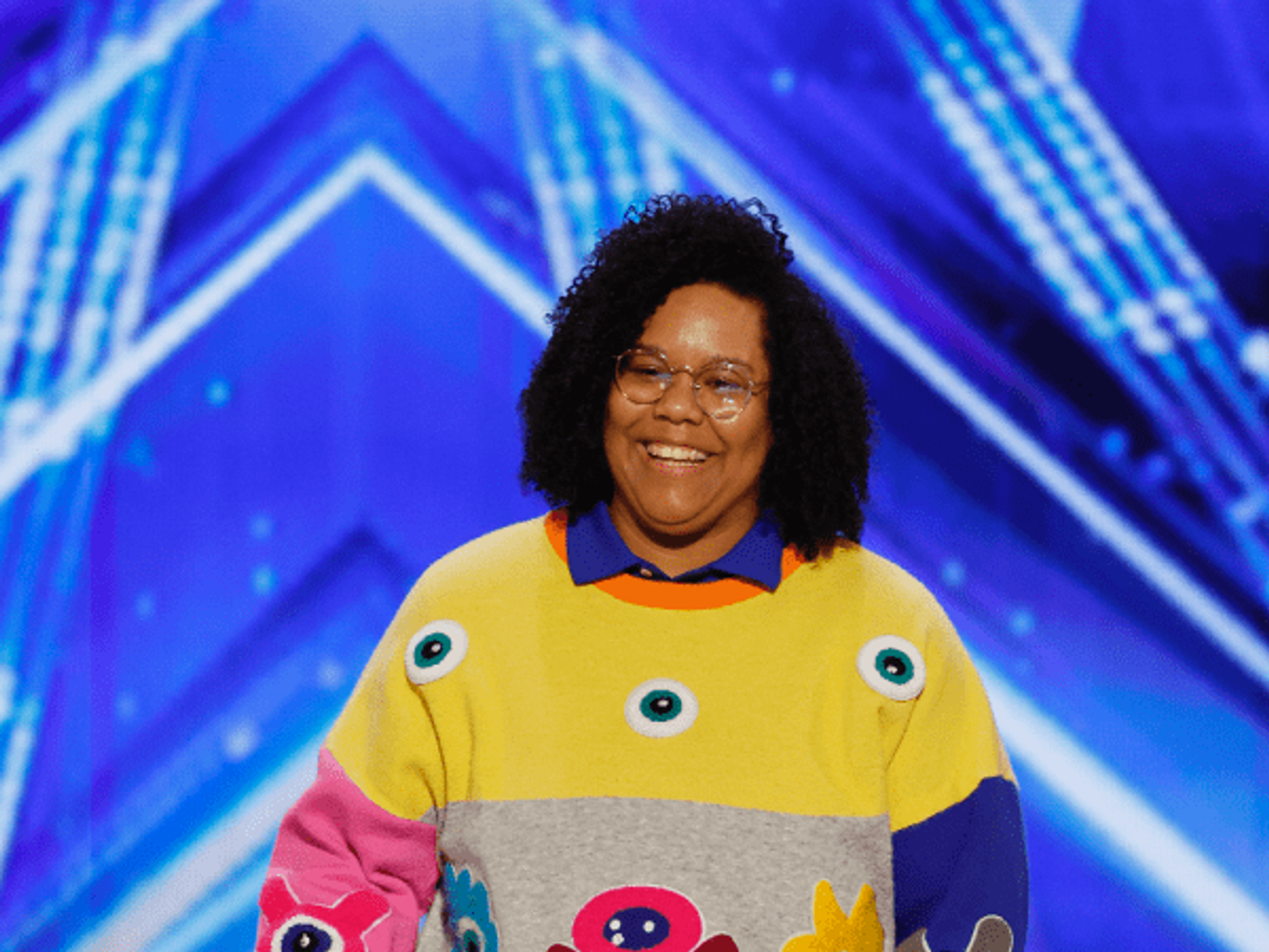 A smiling performer, Mama Duke, stands on stage, wearing a colorful sweater with cartoon monster designs, dark jeans, and pastel sneakers, holding a microphone against a backdrop of vibrant stage lights.