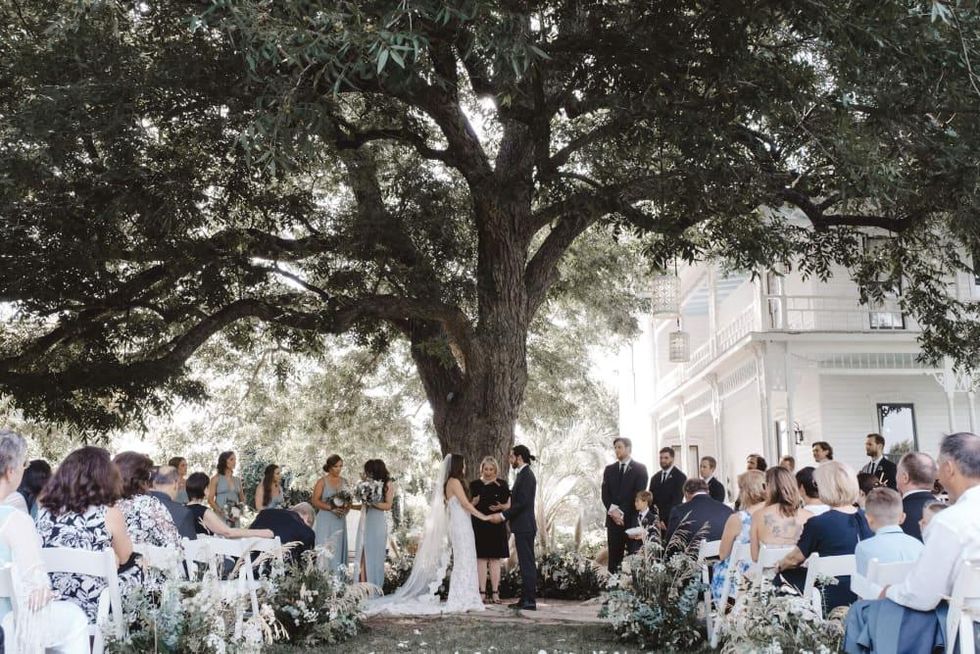 A wedding ceremony is underway under a huge oak tree, next to a white house.