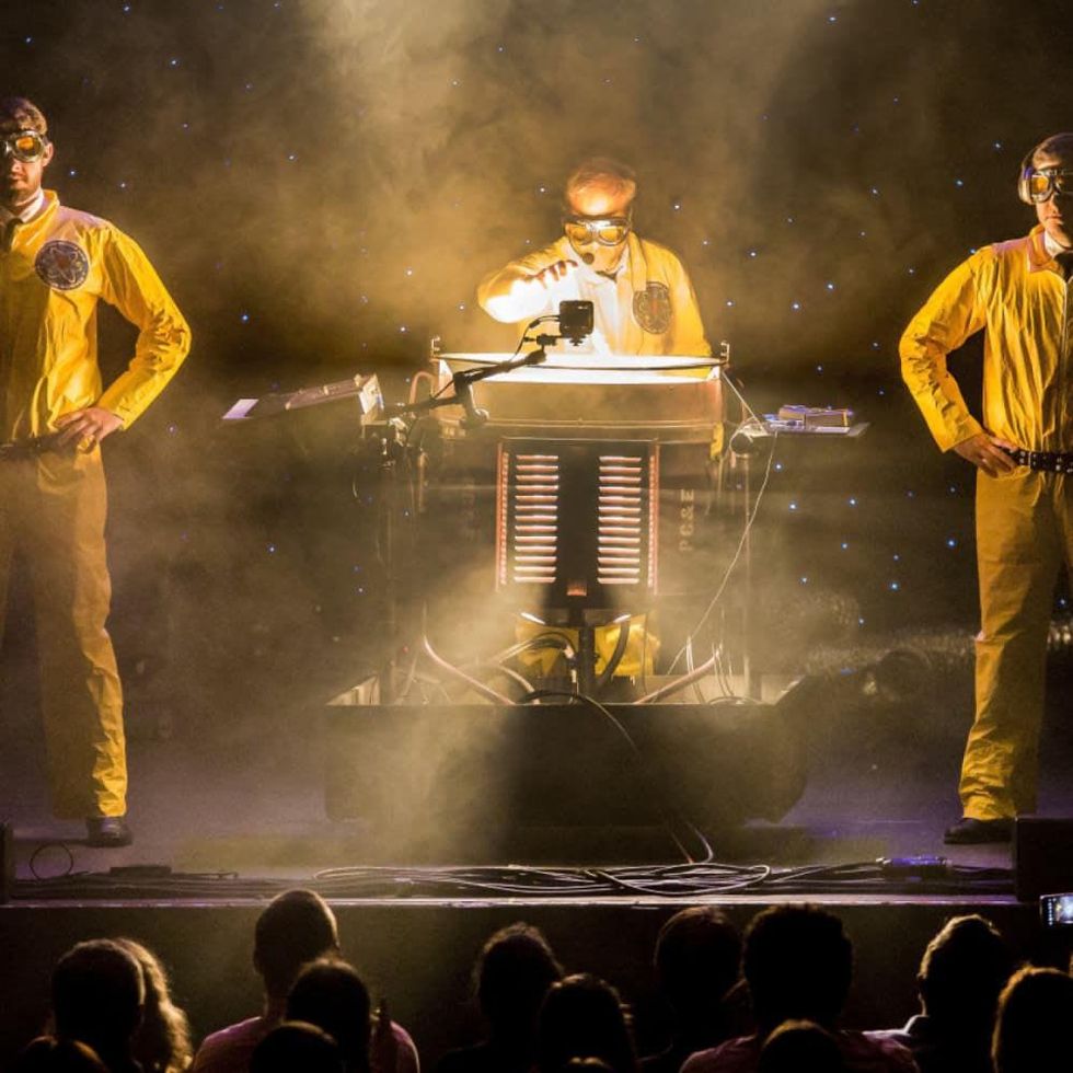 Alton Brown and two assistants stand in yellow hazmat suits around a mysterious contraption.